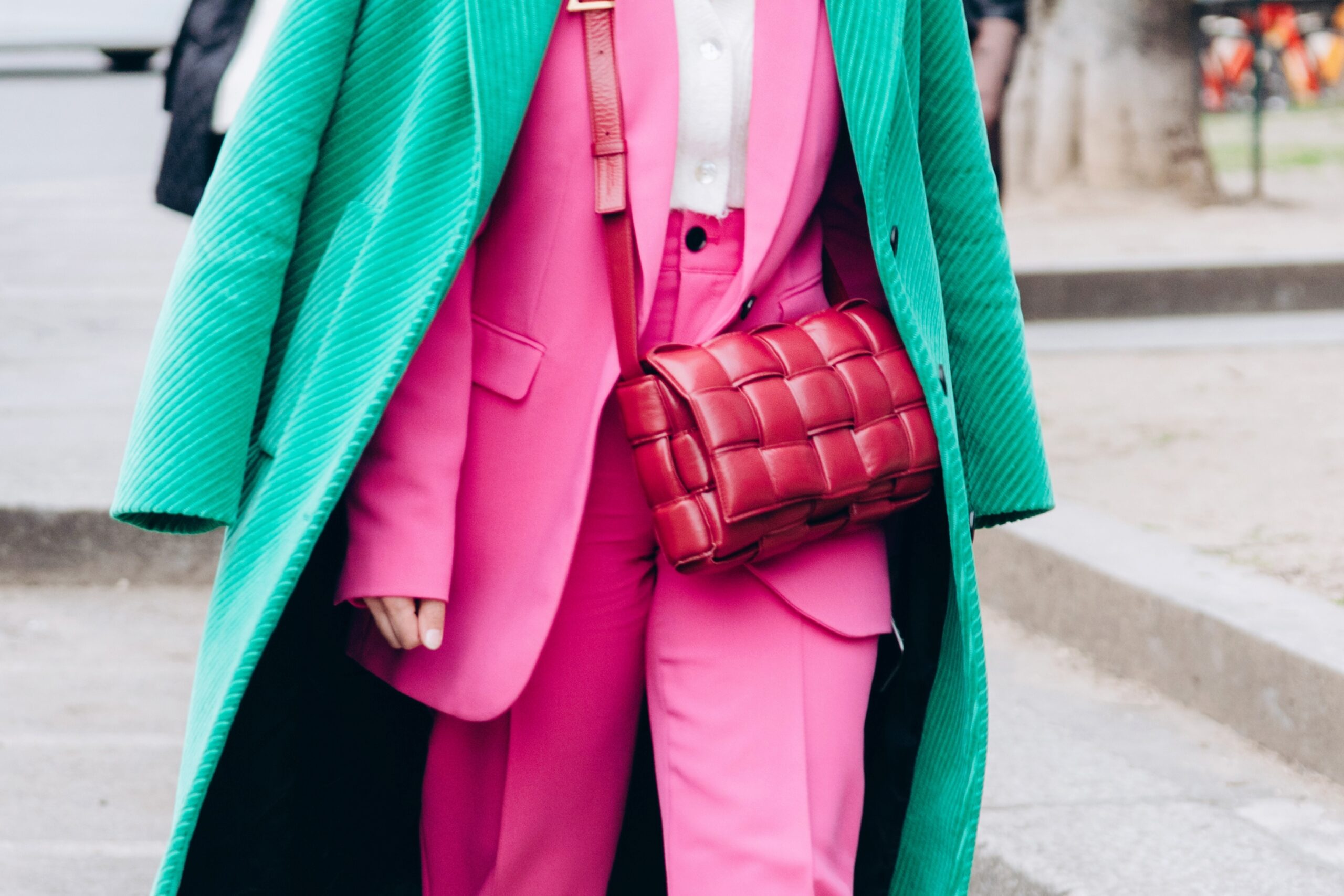 Person wearing a vibrant pink suit paired with a green textured overcoat and carrying a signature woven red Bottega Veneta bag, photographed on a city street.